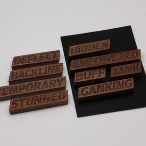 May include: A collection of small, rectangular wooden tiles featuring engraved gaming terminology. The words include DEFLECT, BACKLINE, TEMPORARY, STUNNED, HIDDEN, EMPOWERED, BUFF, TANK, and GANKING. Some tiles are arranged on a black rectangular card.