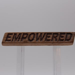 Può includere: Una barra rettangolare in legno con venature marrone scuro, caratterizzata dalla parola EMPOWERED incisa in lettere maiuscole sulla superficie anteriore. Il pezzo ha una finitura liscia ed è esposto orizzontalmente, mettendo in risalto la texture naturale.