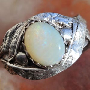May include: A silver ring featuring an oval, iridescent opal gemstone. The ring's setting has a textured, leaf-like design, with a bezel that securely holds the opal. The ring's design is organic and nature-inspired.