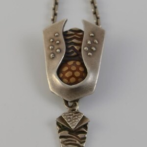 May include: Silver-toned pendant necklace with a unique, abstract design. The pendant features a central brown, textured element, framed by a silver metal structure with small, raised details. A smaller, textured silver element hangs below.