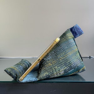 May include: A fabric phone stand in a triangular shape, with a blue and green patterned design. A gold-colored phone is resting on the stand. The stand is made of a soft, textured material.