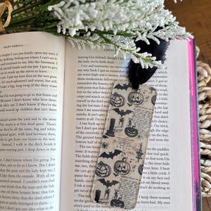 May include: A decorative bookmark featuring a Halloween theme. The bookmark has a black ribbon and is decorated with pumpkins, bats, and other spooky elements. It is placed in an open book with white pages.