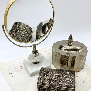 May include: A silver vanity set with a round mirror on a stand, a round silver trinket box with a lid, and a rectangular silver trinket box with an ornate design.