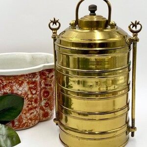 May include: A vintage brass tiered food carrier with a handle and decorative accents. The carrier is cylindrical and has multiple stacked compartments. A floral patterned ceramic container is next to the carrier.