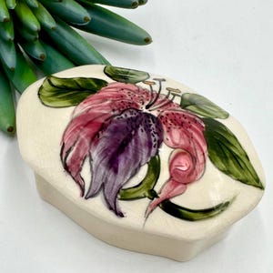 May include: Cream-colored ceramic trinket box with a hand-painted floral design. The lid features a large, stylized flower in shades of pink and purple, with green leaves. The box has a rectangular shape.