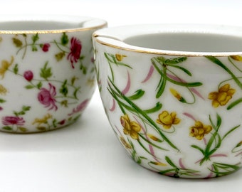 May include: Two white ceramic ashtrays with floral patterns. One features pink and yellow flowers with green leaves, the other has yellow flowers and green leaves with pink accents. Both have gold rims.