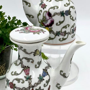May include: Two white ceramic pouring pitchers with floral and butterfly designs. The pitchers have lids and are decorated with green, yellow, pink, blue, and red colors.