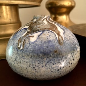 May include: A small, round ceramic vase with a blue, white, and brown marbled glaze. The vase has a speckled texture and a slightly uneven shape.
