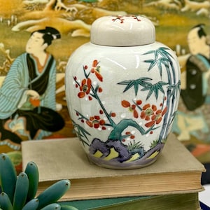 May include: A cream-colored ceramic ginger jar with a lid, featuring hand-painted red flowers and green bamboo. The jar is decorated with a crackle glaze and sits on top of a stack of books. The background features an Asian-inspired painting.