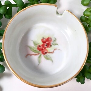 May include: A white porcelain ashtray with a gold rim and a hand-painted design of red berries on a green branch.