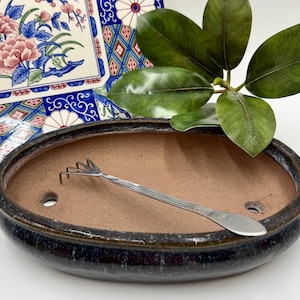 May include: An oval-shaped, dark blue ceramic bonsai pot with a brown interior and two drainage holes. A small, silver metal rake rests inside the pot. A decorative floral plate and green leaves are in the background.