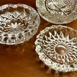 May include: Three clear glass ashtrays with a cut glass design. The ashtrays are round and have a raised lip around the edge.