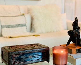 May include: A decorative, antique black and red wooden box with intricate carvings sits on a glass table. A lit, brown-tinted glass candle and a bronze sculpture of a figure riding a fish are also on the table. A white sofa with pillows is in the background.