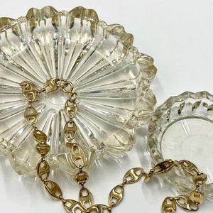 May include: Two clear glass ashtrays with a gold chain link necklace resting on top. The larger ashtray has a scalloped edge and a textured bottom. The smaller ashtray has a faceted edge.