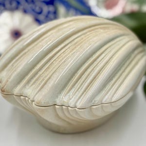 May include: A cream-colored, ceramic seashell-shaped trinket box. The box has a ridged texture and a glossy finish. The lid fits snugly onto the base, creating a secure closure. The box is a decorative item.