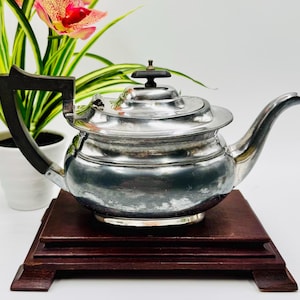 May include: A silver-colored teapot with a dark wooden handle and lid knob sits on a dark wooden stand. The teapot has a curved spout and a round body. A plant with green and yellow leaves and a pink and yellow flower is in the background.