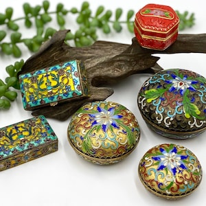 May include: A collection of ornate, antique cloisonné boxes in various shapes and sizes. The boxes feature intricate floral and geometric designs in vibrant colors like red, yellow, blue, and green, with gold accents. The boxes are displayed on a wooden surface.