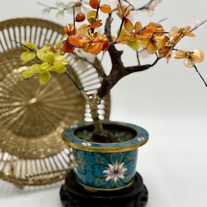 May include: A decorative bonsai tree with orange, yellow, and white flower-shaped accents. The tree is in a turquoise pot with a floral design, set on a black stand. A gold-colored, intricately designed plate is in the background.