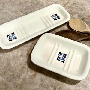 May include: Two white ceramic soap dishes with raised ridges and a central square graphic featuring a blue and white floral motif on a black background, accompanied by one light-colored wooden spoon and one dark-colored wooden spoon.