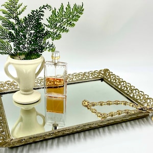 May include: A gold mirrored tray with a decorative filigree border. A clear glass bottle of perfume sits on the tray, along with a gold chain necklace. A white ceramic vase with two handles holds a sprig of green ferns.