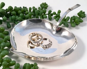 May include: A silver leaf-shaped trinket dish with a handle, featuring a smooth, reflective surface. The dish holds several rings, including a diamond ring.
