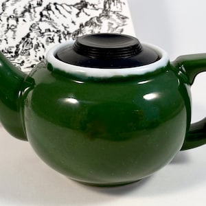 May include: A small, green ceramic teapot with a black lid and a white rim. The teapot has a round body and a curved spout.