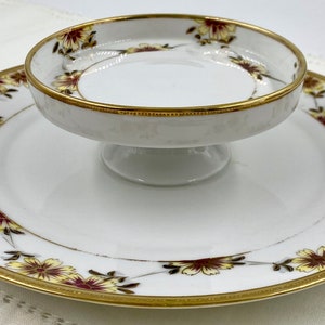 May include: A white porcelain serving dish with a gold rim and floral design. The dish has a smaller, matching dish on a pedestal in the center.