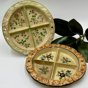 May include: Two vintage divided serving dishes. The round dish has a cream-colored base with green and pink floral designs. The oval dish has a tan base with floral designs. Both dishes have a decorative border.