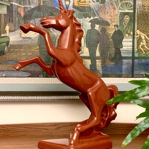 May include: A brown ceramic unicorn figurine with a single horn, standing on its hind legs with its front legs raised. The figurine is on a brown wooden surface.