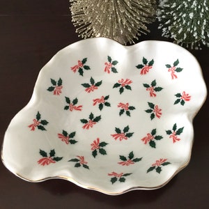 May include: White ceramic dish with a scalloped edge and a gold rim. The dish is decorated with a repeating pattern of green holly leaves and red bows.