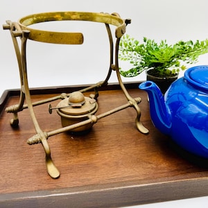 May include: A vintage brass tea warmer with a circular top and a small burner. A bright blue teapot sits nearby on a wooden tray, with a small potted plant in the background. The tea warmer is an antique style.