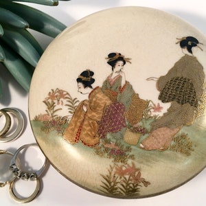 May include: A round, cream-colored trinket box with a lid featuring a hand-painted scene of three women in traditional Japanese clothing. The women are surrounded by flowers and greenery, and the box is decorated with gold accents.
