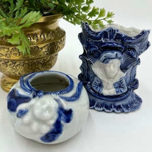 May include: Three decorative ceramic planters, one gold and two blue and white. The blue and white planters feature floral designs and one has a portrait of a person.