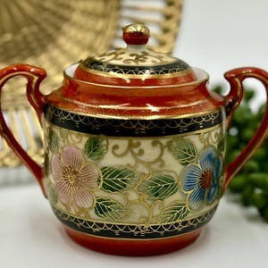 May include: A red and white porcelain sugar bowl with a lid. The bowl has a floral design with gold accents and a black band around the top and bottom. The lid has a gold knob.