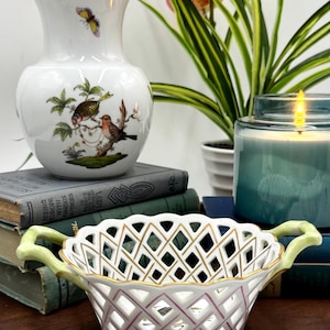 May include: A white porcelain vase with a gold rim and a butterfly and bird design. A white, openwork porcelain basket with gold trim and green handles. A blue candle and books are also visible.