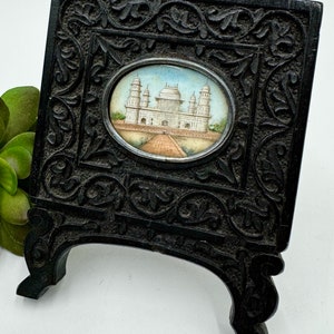 May include: A black wooden frame with intricate carvings, holding a miniature painting of the Taj Mahal. The frame has a stand on the back, allowing it to be displayed upright.