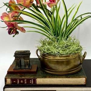 May include: A brass planter with green moss and a small replica of the Arc de Triomphe on a stack of books. The books have titles including "A Voyage to the South Seas" and "The Limited Editions Club".