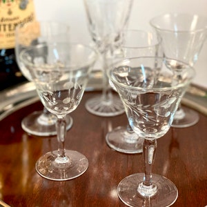 May include: Five clear glass stemmed drinking glasses with etched designs on a wooden surface. The glasses are arranged in a circle, with the stems pointing outwards.
