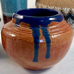 May include: A small, round, terracotta-colored ceramic pot with a blue glaze dripping down the sides. The pot has a smooth, glossy finish.