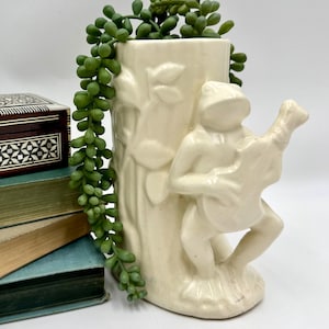 May include: A cream-colored ceramic vase shaped like a tree trunk with a sculpted frog playing a guitar attached to the front. The vase is filled with a trailing green succulent plant, creating a decorative piece with a smooth, glossy finish.