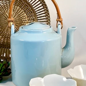 May include: A light blue ceramic teapot with a brown woven handle. The teapot is sitting on a white surface with two small white bowls with yellow rims.