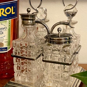 May include: A silver-plated, three-bottle cruet set with a wooden handle. The cruets are made of clear glass with a textured, square pattern. The set is on a silver-plated base.