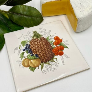 May include: A white ceramic tile with a pineapple, cherries, blackberries, and two peaches in a floral design. The tile has a white background with gray and green foliage.