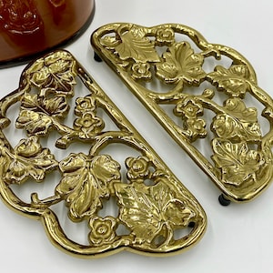 May include: A set of two gold-tone metal hot pads with a leaf and floral design.