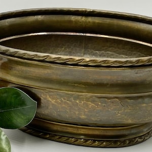 Puede incluir: Un macetero ovalado de latón con acabado martillado y un diseño de cuerda alrededor de los bordes superior e inferior. El macetero tiene dos asas de cabeza de león con anillos.