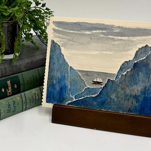 May include: Watercolor painting of a seascape with a sailboat between blue cliffs under a gray sky. The artwork is displayed on a wooden stand, with a stack of vintage books and a small potted plant in the background.