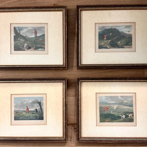 May include: A set of four framed prints depicting outdoor hunting scenes with figures in red coats and hunting dogs. Each print is housed in a dark brown wooden frame with a cream-colored mat, showcasing detailed landscape illustrations.