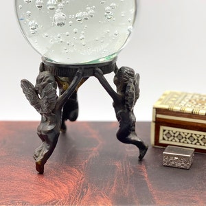 May include: A clear glass orb with air bubbles, resting on a bronze-colored stand supported by three cherub figures. A small decorative box and a book are also visible.
