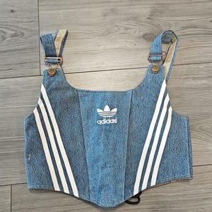 Peut inclure: Un haut court style corset en jean bleu arborant le logo trèfle Adidas blanc centré sur la poitrine. Le vêtement comprend deux bretelles avec des boucles métalliques et trois bandes verticales blanches sur chaque côté du bustier.