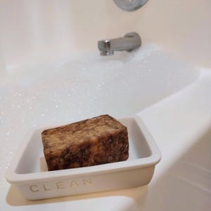 May include: A white ceramic soap dish with the word "CLEAN" embossed on the front. A brown bar of soap is sitting inside the dish. The dish is in a white bathtub with bubbles.